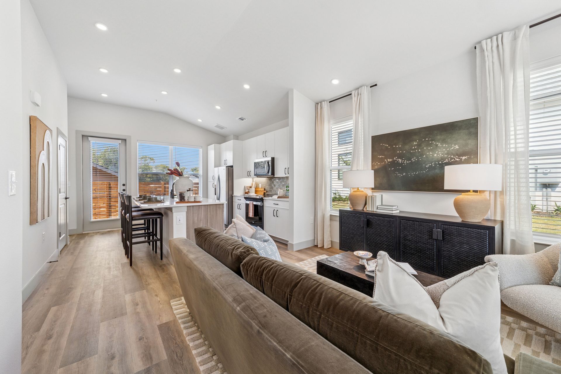 Open-concept living space with kitchen, living room, and dining area; neutral colors, hardwood floors, and large windows.