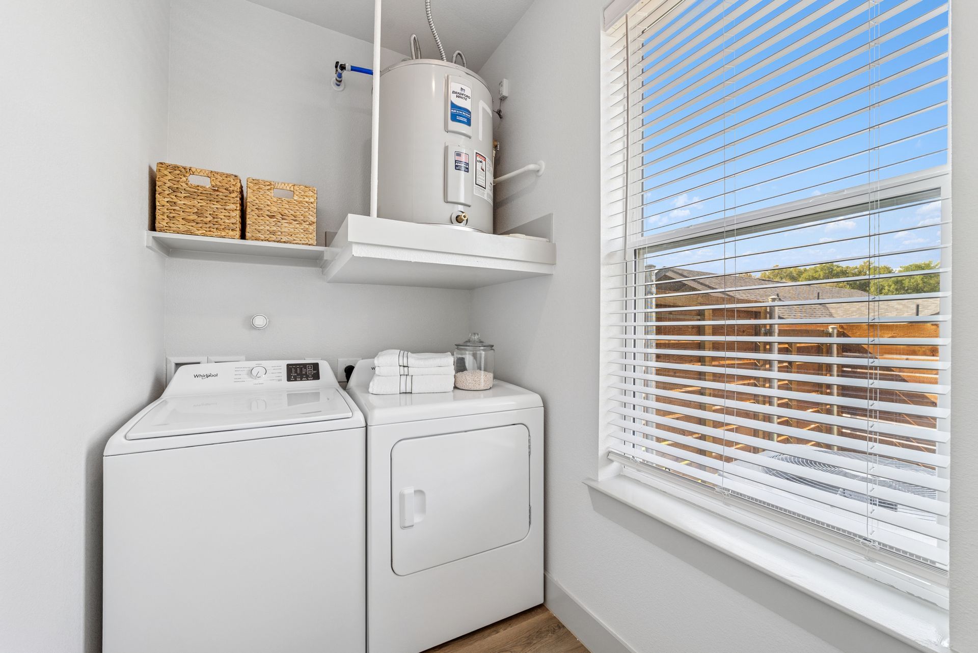 Laundry room with a washer, dryer, water heater, shelf with baskets, and a window with blinds.
