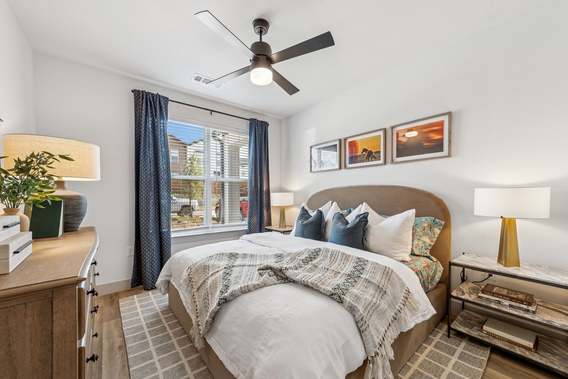 Bedroom with a bed, dresser, window, art, and a ceiling fan; earth tones with blue accents.