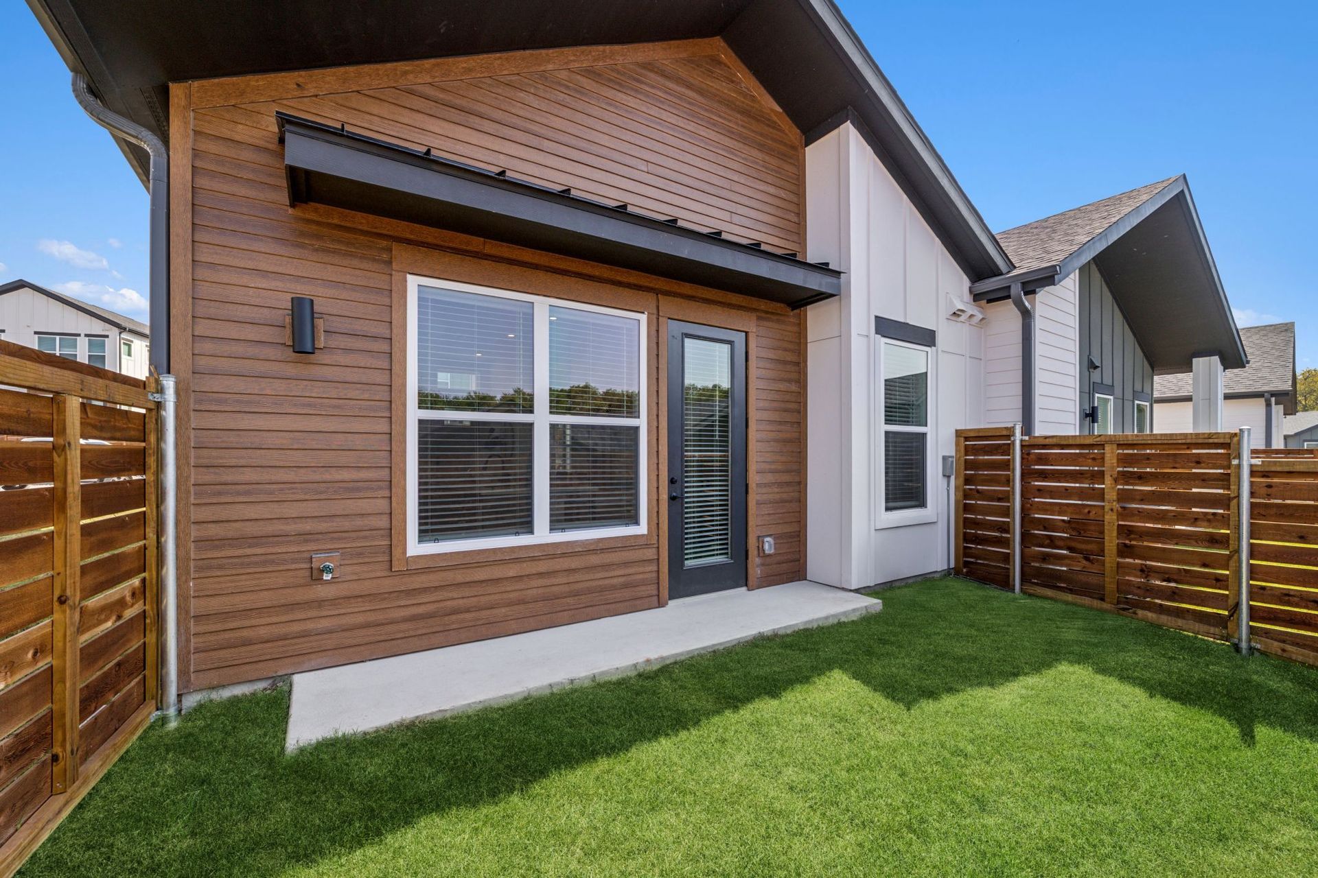Exterior of a modern home with brown siding, windows, and a small patio surrounded by a wooden fence and green grass.