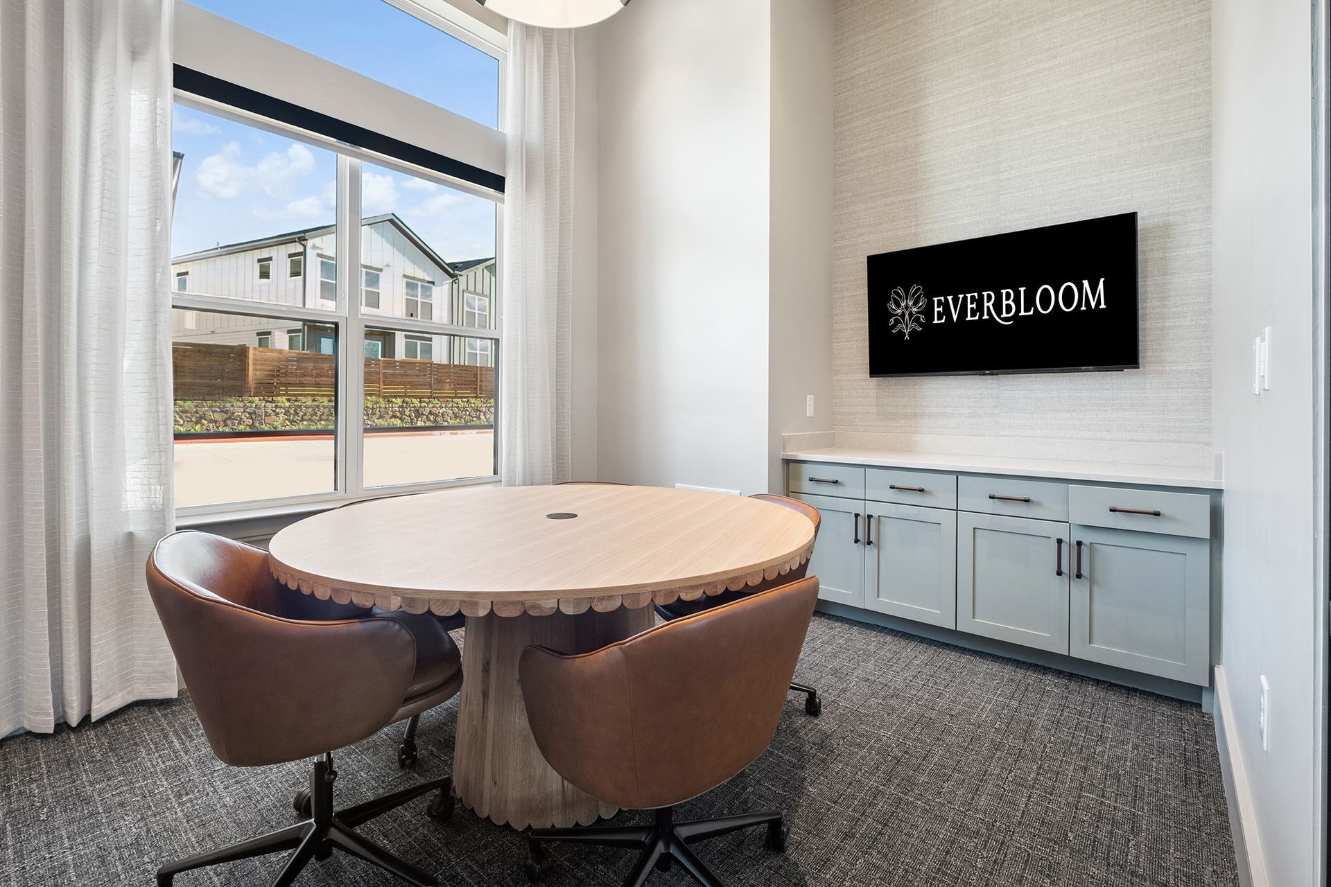 A modern meeting room with a round wooden table, chairs, and a TV; a large window reveals the outdoors.