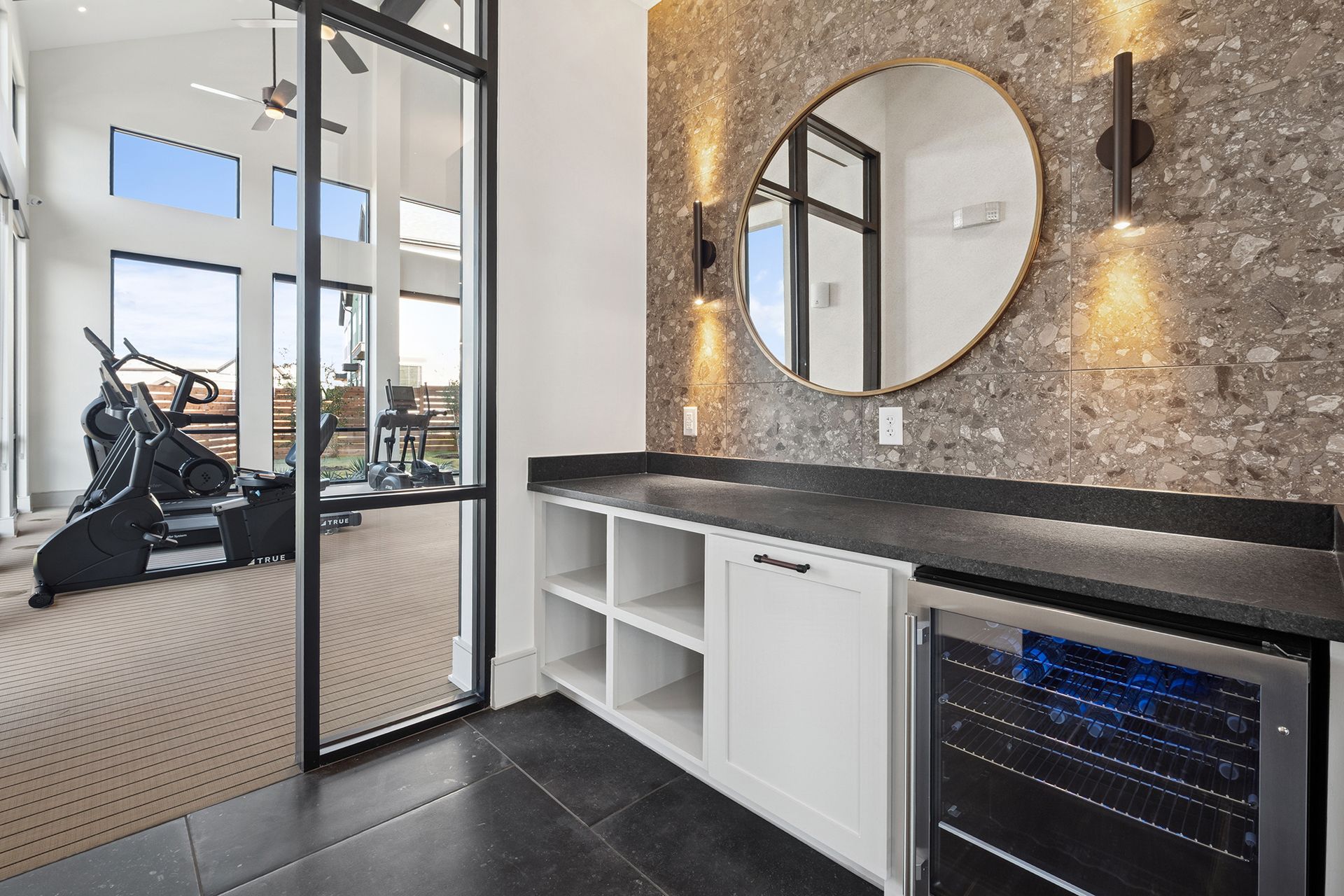 Fitness room with equipment visible through a glass door, built-in bar with a mirror, and a wine fridge.