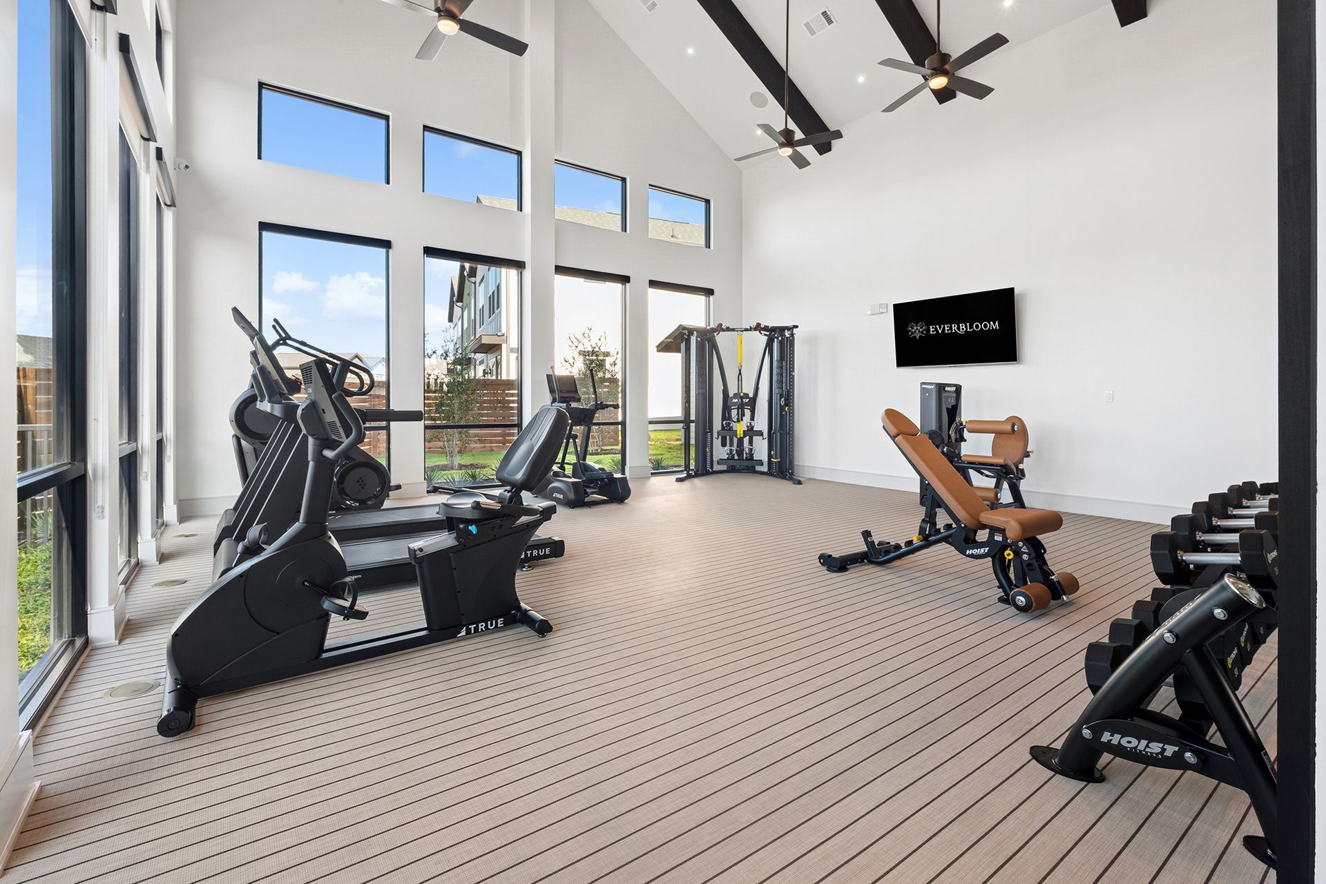Gym with cardio equipment, weight bench, and free weights, under tall windows.