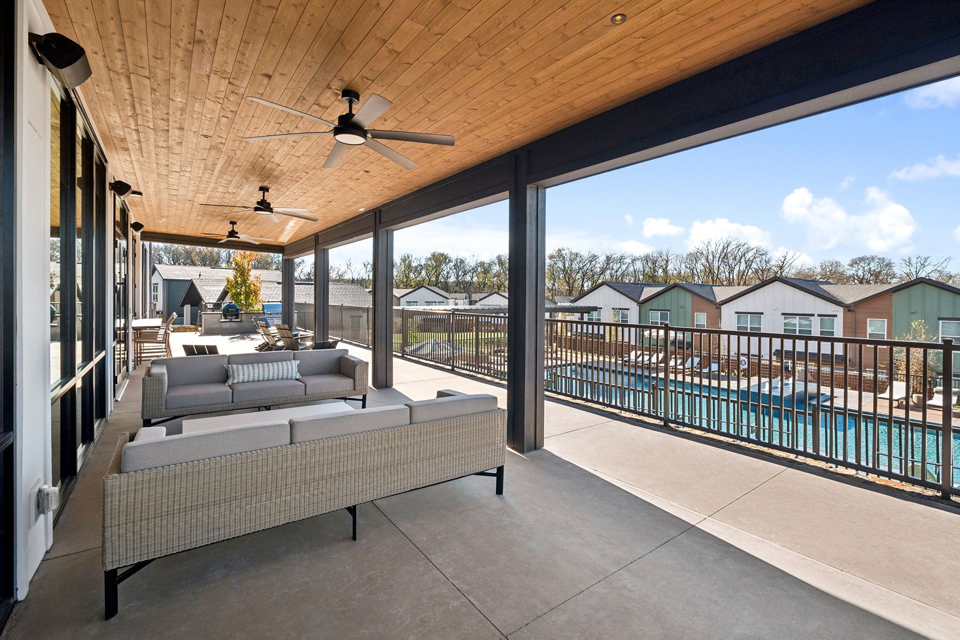 Covered patio overlooking a pool and townhouses; gray sofa, wood ceiling, and black railings.