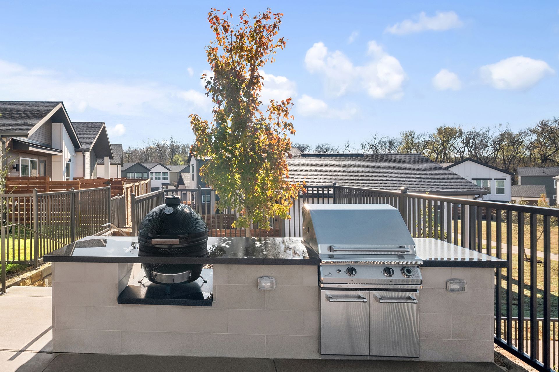 Outdoor kitchen with built-in grill and Big Green Egg, black countertop, neutral colored base, black railing, blue sky.