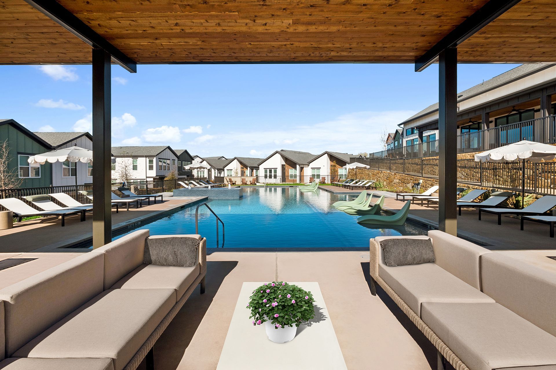 Outdoor lounge area with pool view: tan sofas, blue water, buildings, sunny sky.
