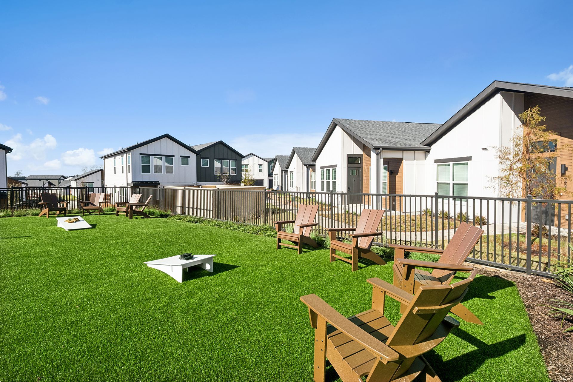 Lawn area with Adirondack chairs and cornhole game, surrounded by modern homes under a blue sky.