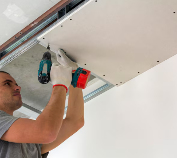 Persona instalando paneles de yeso en un techo con un taladro; lleva guantes.