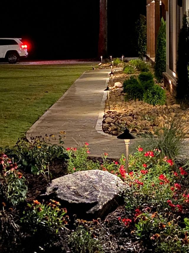 Lit walkway beside landscaped garden beds at night, with a car in the distance.