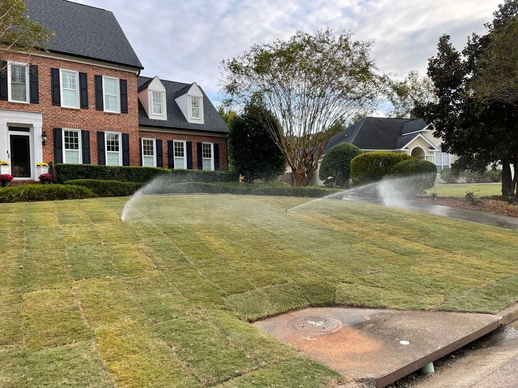 Lawn sprinklers watering a suburban front yard beside a brick house on a cloudy day