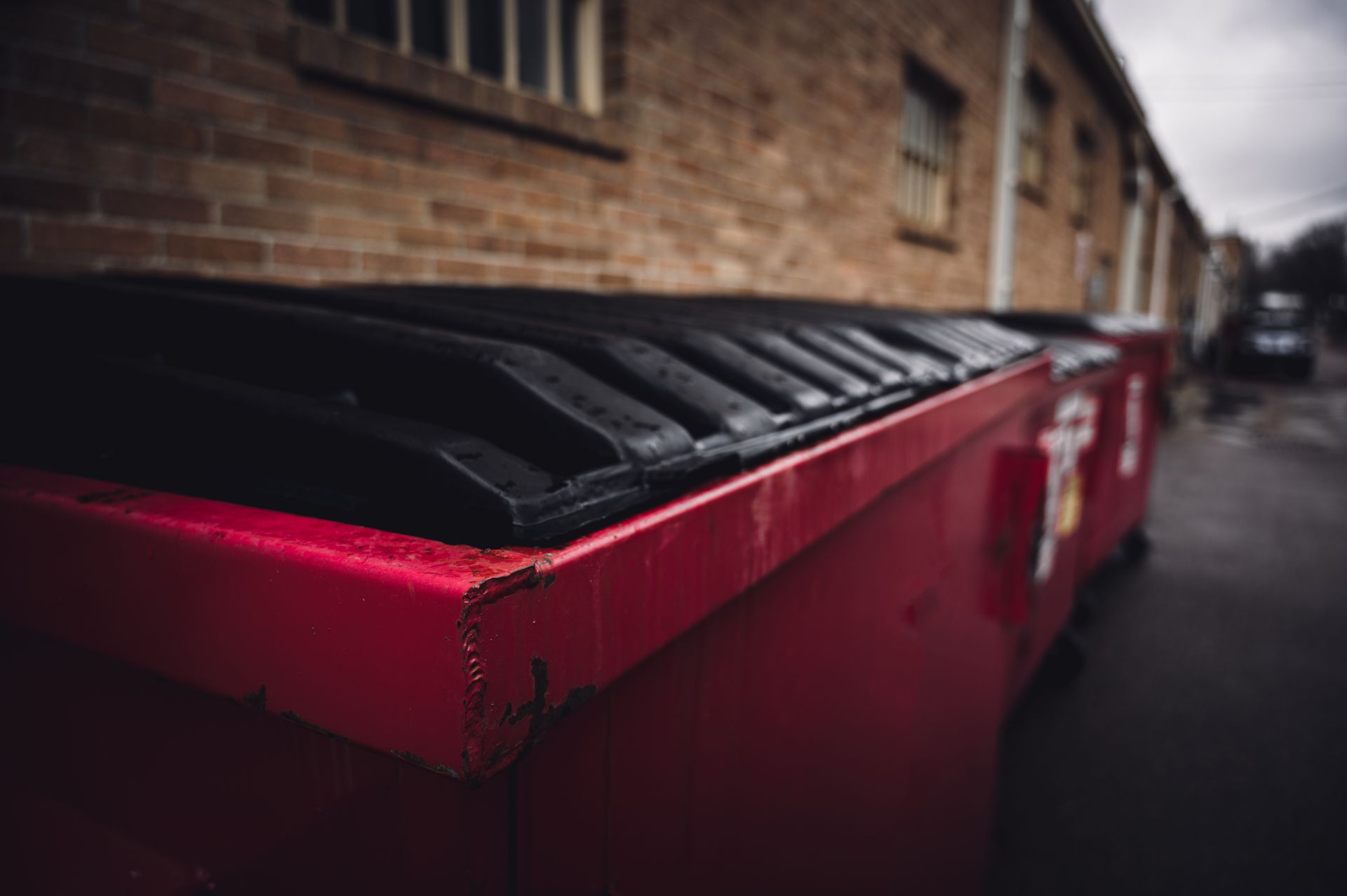 Red dumpster, black lid, brick building in background on an overcast day.