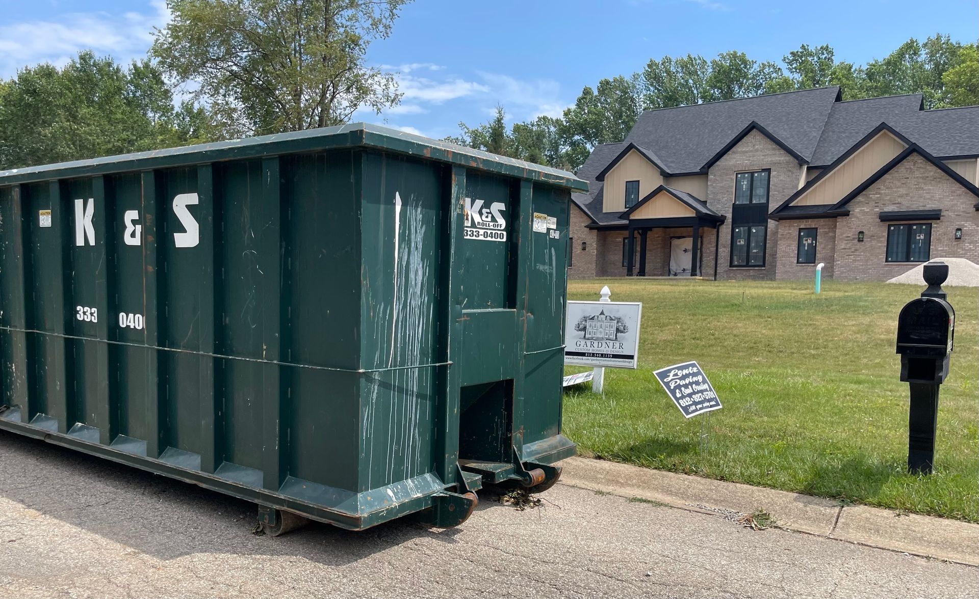 A large green dumpster is parked in front of a large house.