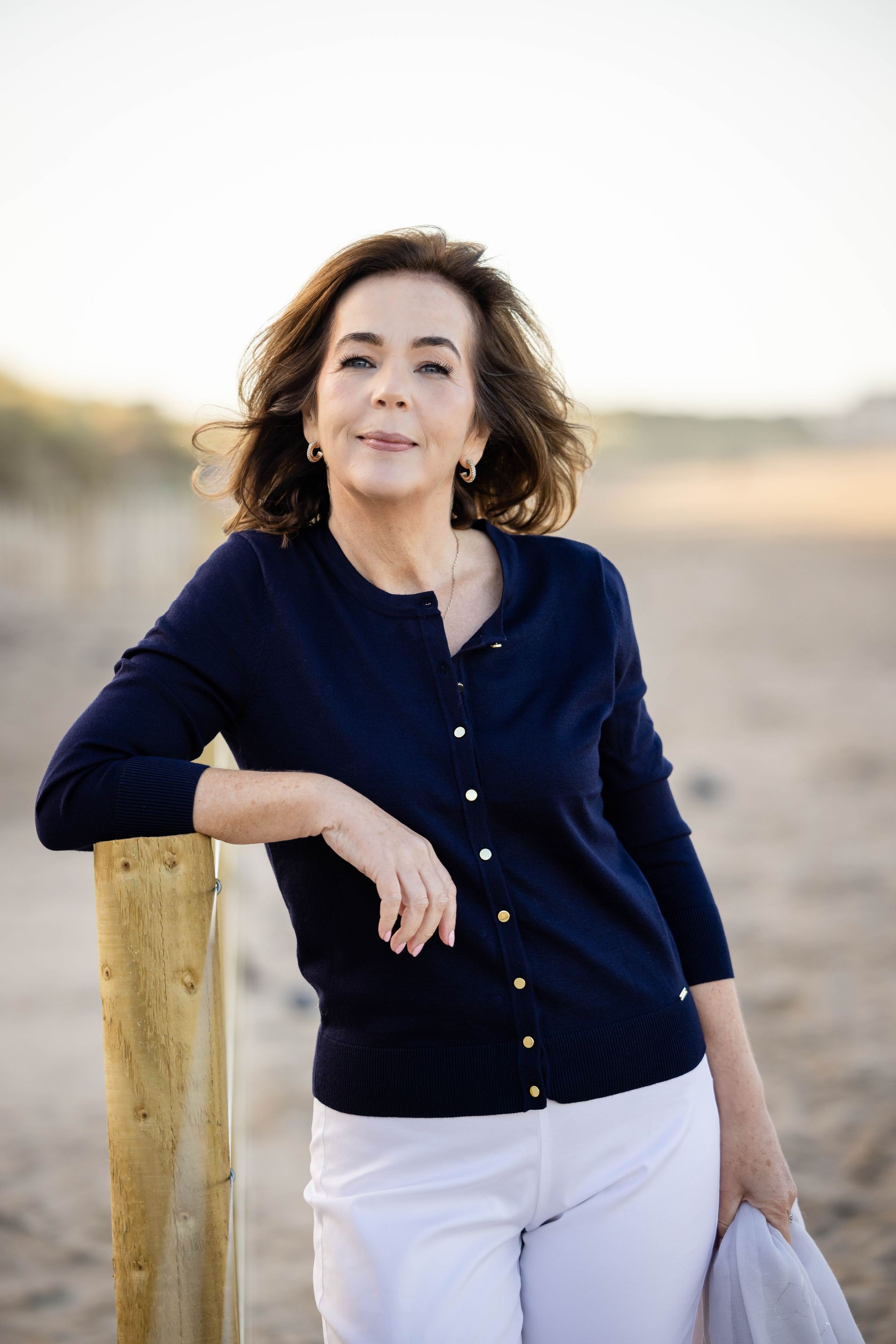 A woman in a blue cardigan and white pants is leaning on a wooden post on the beach.
