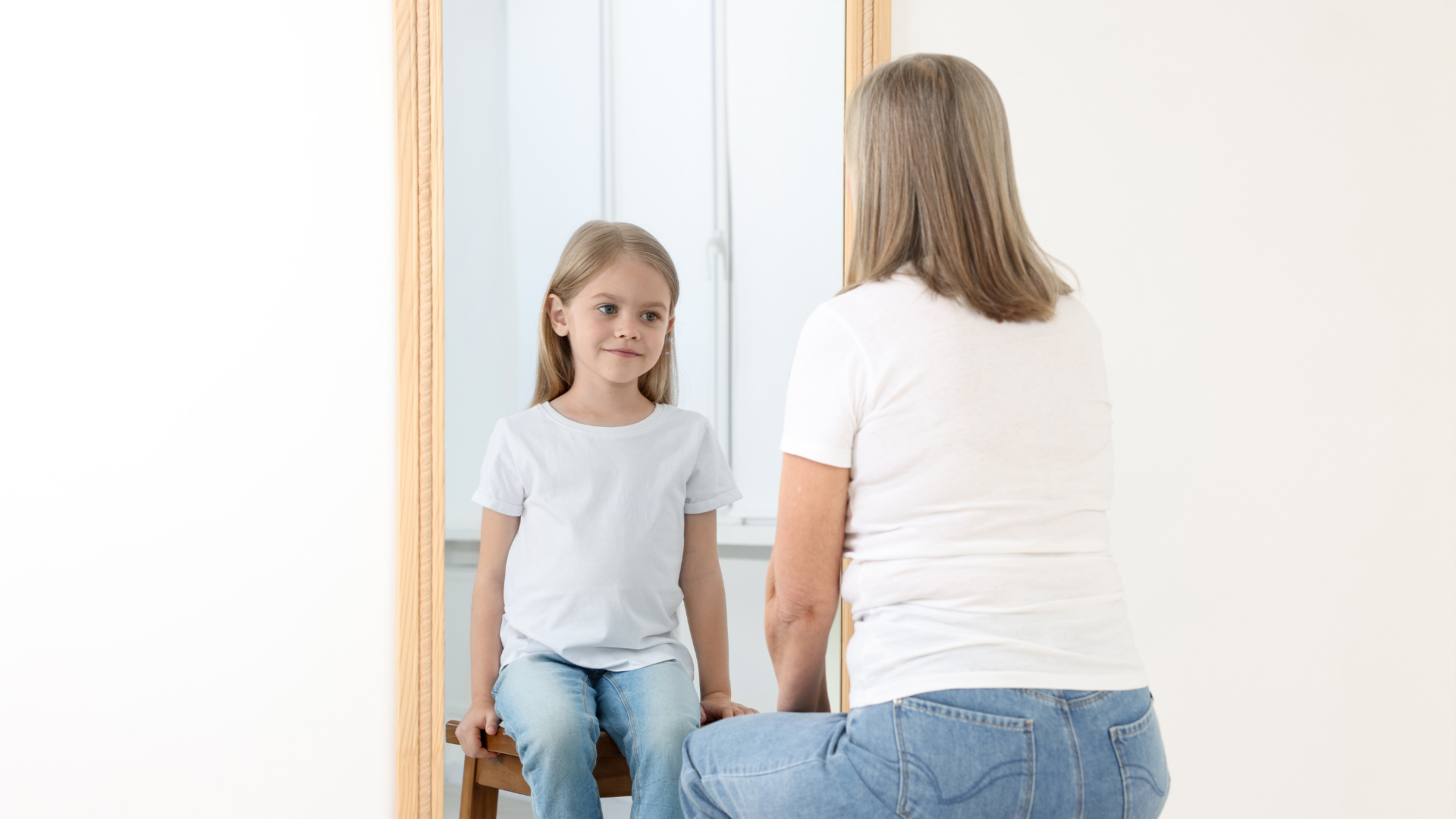 A woman sits facing a mirror. A girl looks at the mirror. Both wear white shirts and jeans, in a white room.