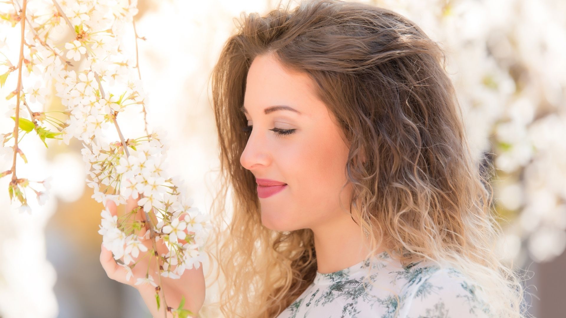 Woman with curly blonde hair smelling white flowers, smiling in the sunlight.