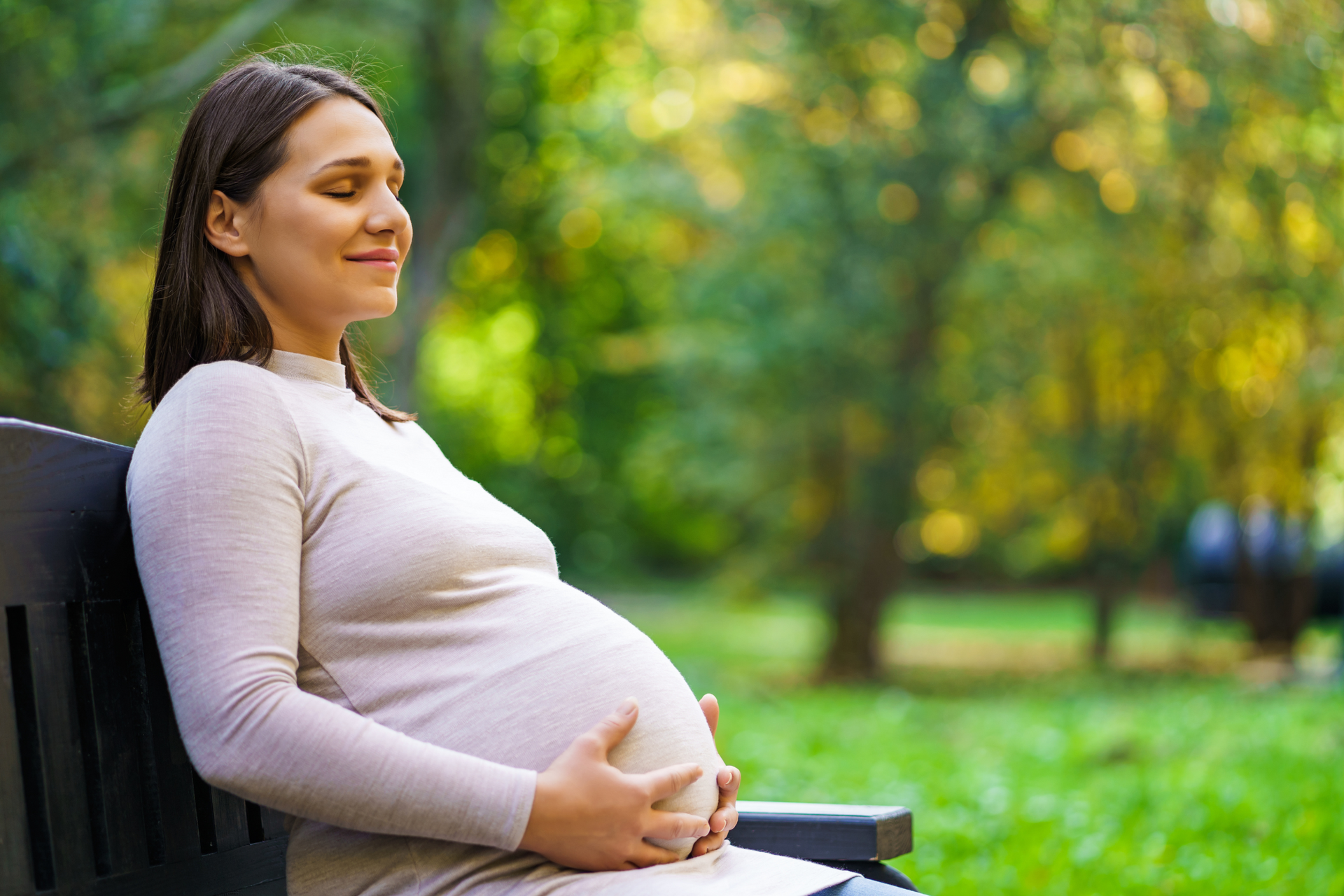 A pregnant woman is sitting on a park bench holding her belly.
