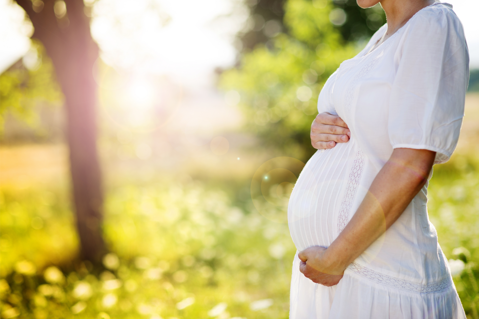A pregnant woman in a white dress is holding her belly in a park.