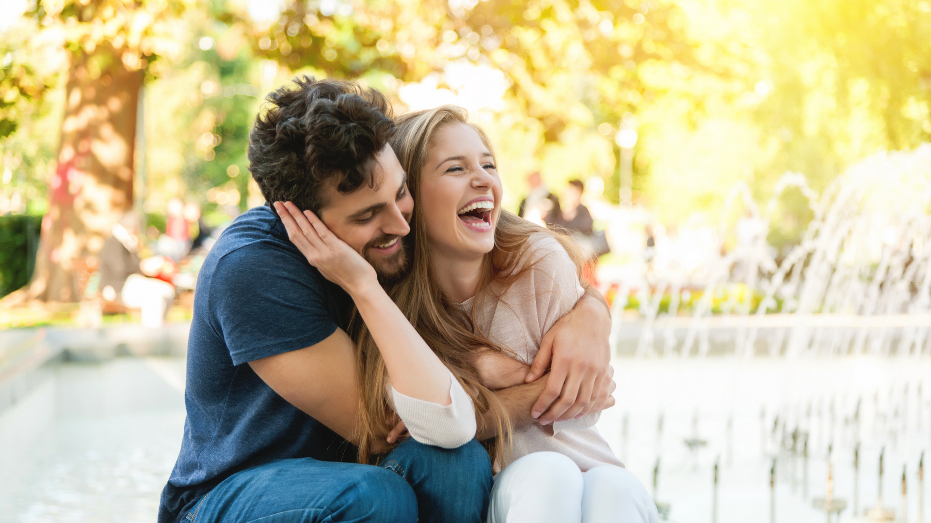 A man and a woman are sitting next to each other in front of a fountain and laughing.