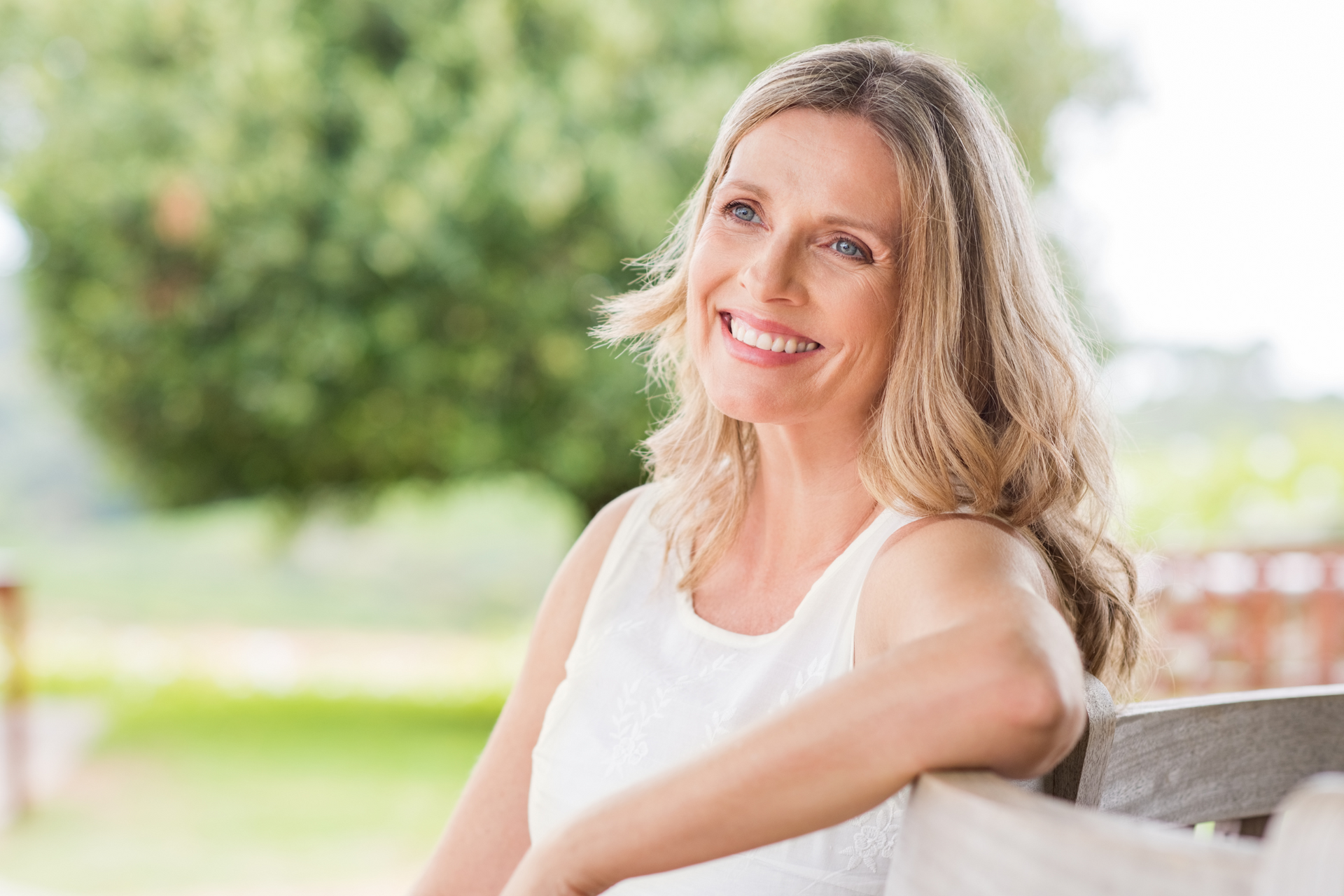 A woman in a white tank top is sitting on a bench and smiling.