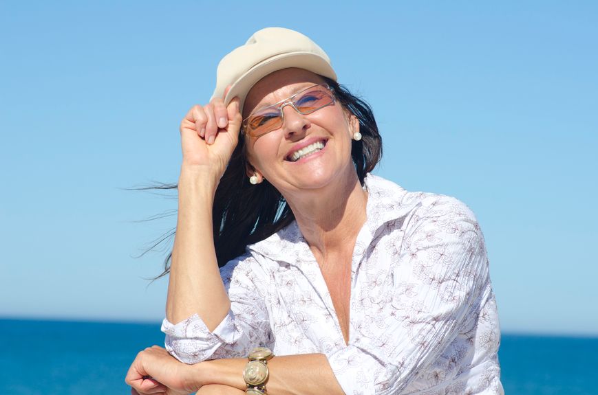 A woman wearing a hat and sunglasses is smiling at the beach.