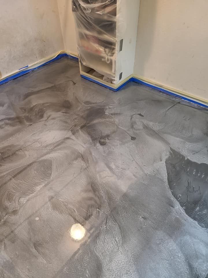 Metallic gray epoxy flooring being installed in a room, edges taped, column wrapped in plastic.