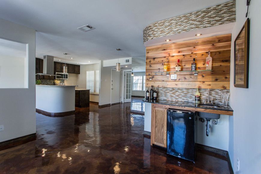 Interior view of home with a bar area and kitchen in the background, featuring wood paneling, small fridge, and dark flooring.