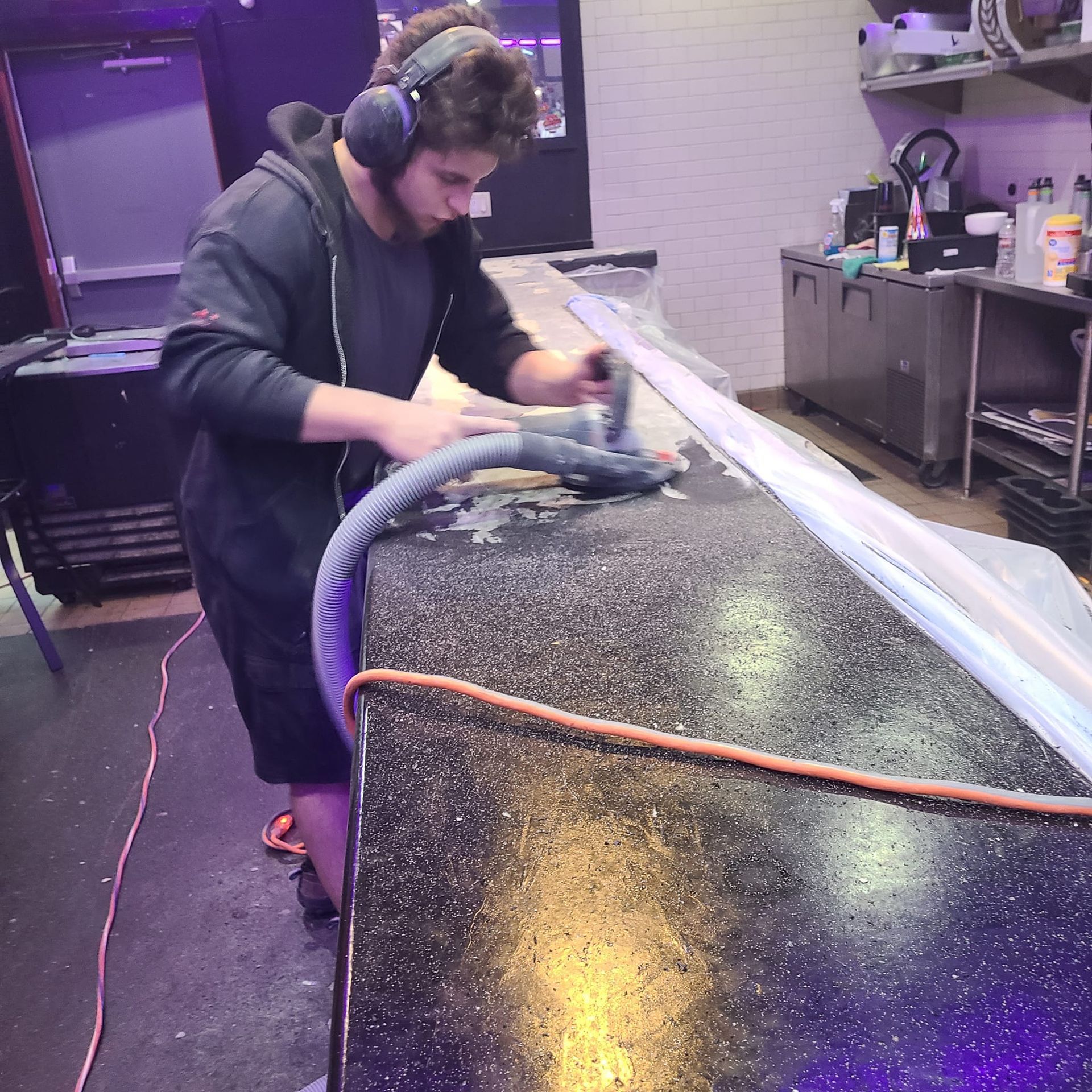 A man wearing earmuffs uses a sander to clean a countertop in a shop.