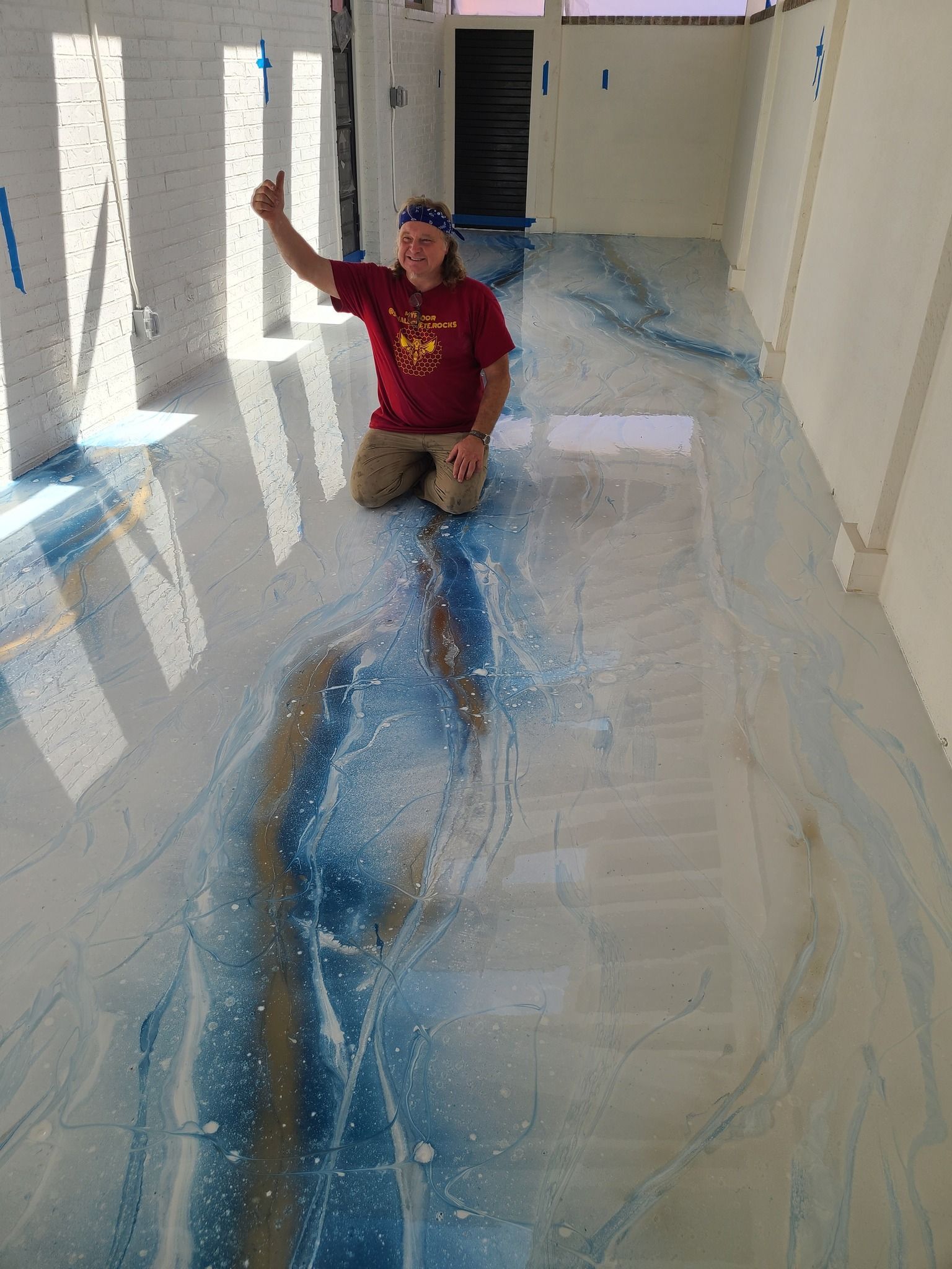 Man kneeling on a blue and white epoxy floor giving a thumbs up, inside a room with white walls.