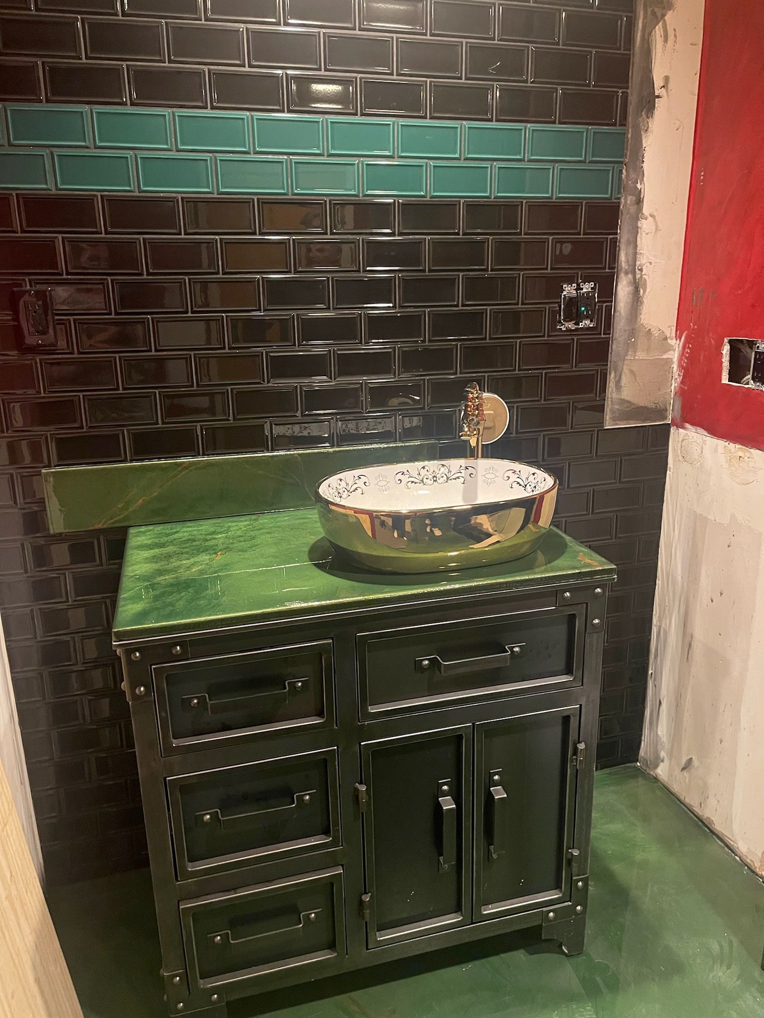 Bathroom with black and teal tiled wall, green countertop and cabinet, gold sink basin.