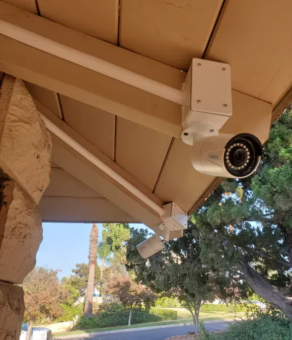 Security camera mounted on a beige building overhang, pointing outward. Another spotlight camera is also mounted nearby.