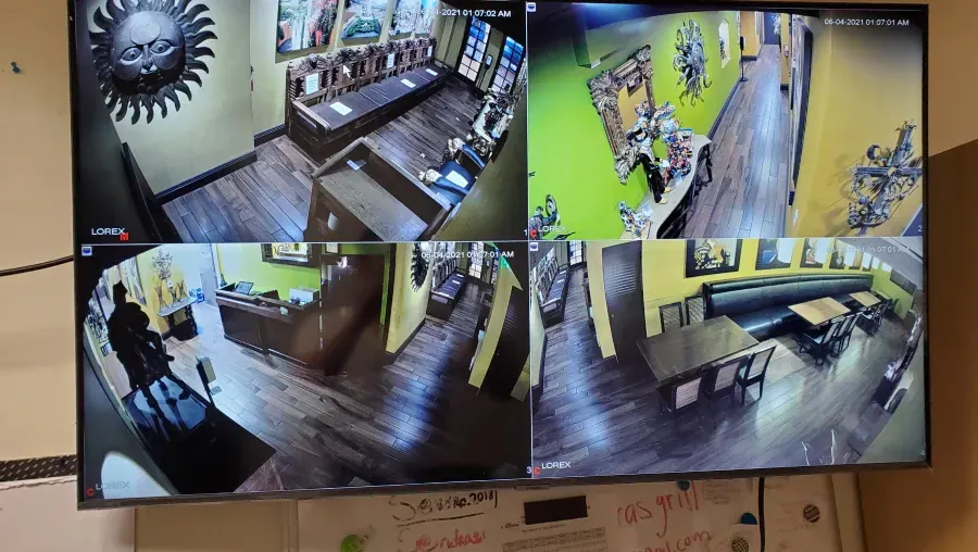 A multi-screen security monitor displaying surveillance footage of a business interior, showing different rooms with artwork, furniture, and an entrance.