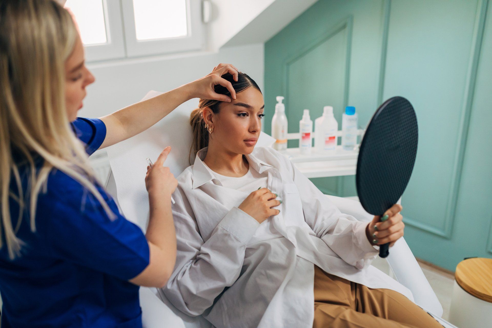 A woman examines her face in a hand mirror while a medical professional points at her forehead.