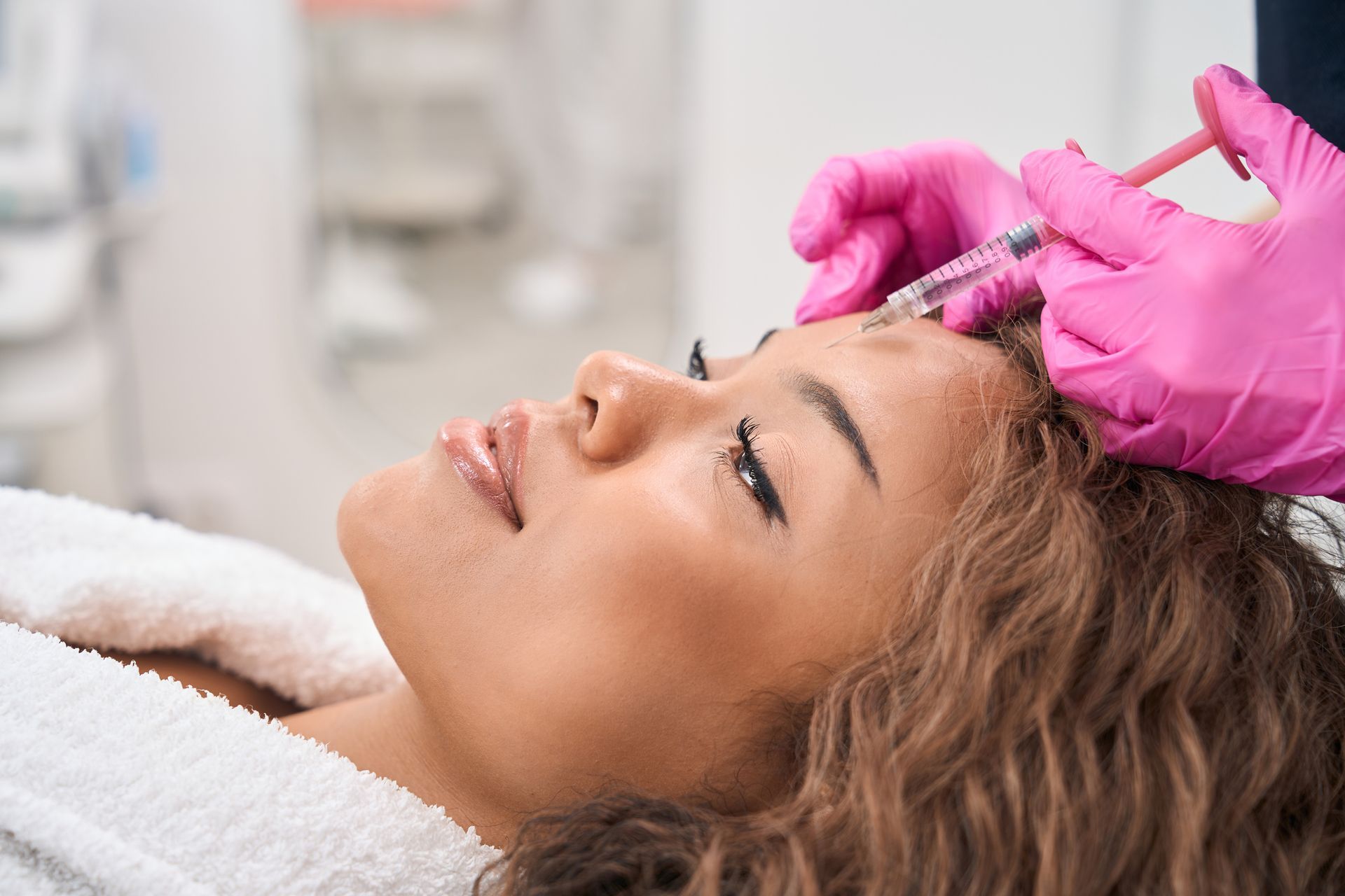 A woman receives cosmetic Botox treatment at a clinic, from an esthetician wearing pink gloves.