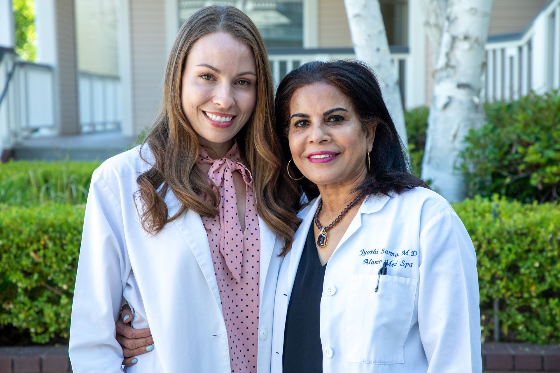 Two women in white lab coats smile outdoors, near greenery.