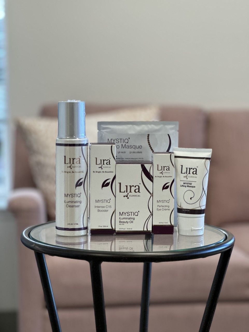 Skincare products on a glass table in front of a pink sofa. Lyra brand products with white and purple labels.