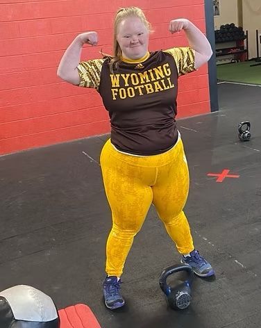 Woman flexes, wearing Wyoming Football shirt and yellow leggings, in gym with kettlebell.