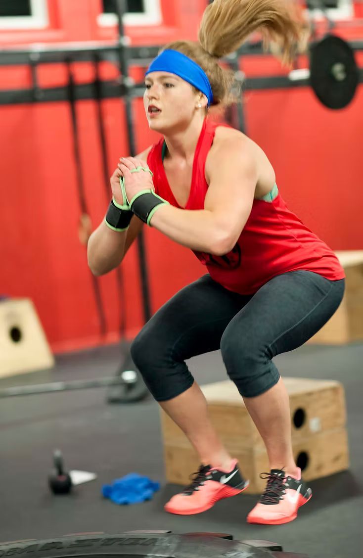 Woman in red tank top and blue headband doing a box jump in a gym.