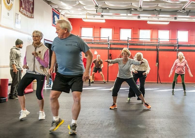People exercising in a gym; several performing side steps, others in the background. Red wall, gray floor.