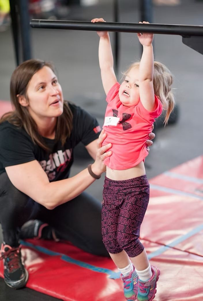 Woman spotting a young child hanging from a bar; indoor gym setting.