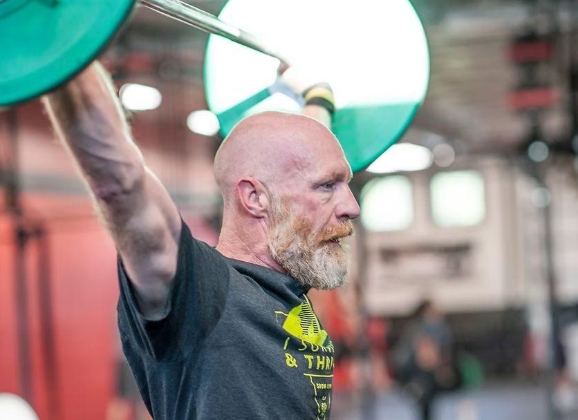 Bald man with a beard lifting a barbell overhead in a gym.