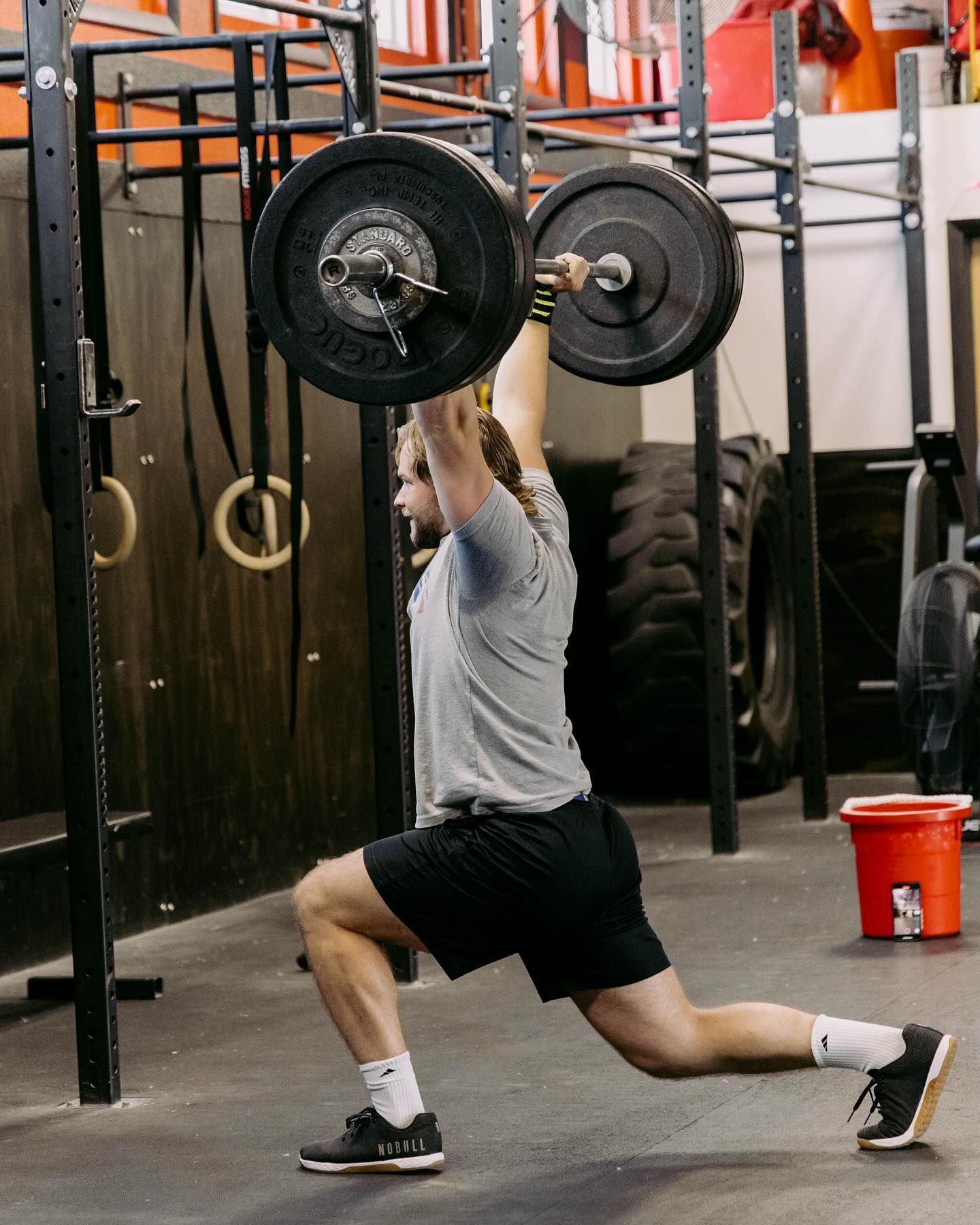 Man lunging with barbell overhead in a gym.