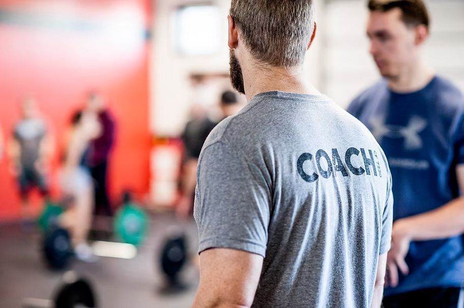 Coach wearing gray shirt watches athletes at a gym; another man stands nearby.