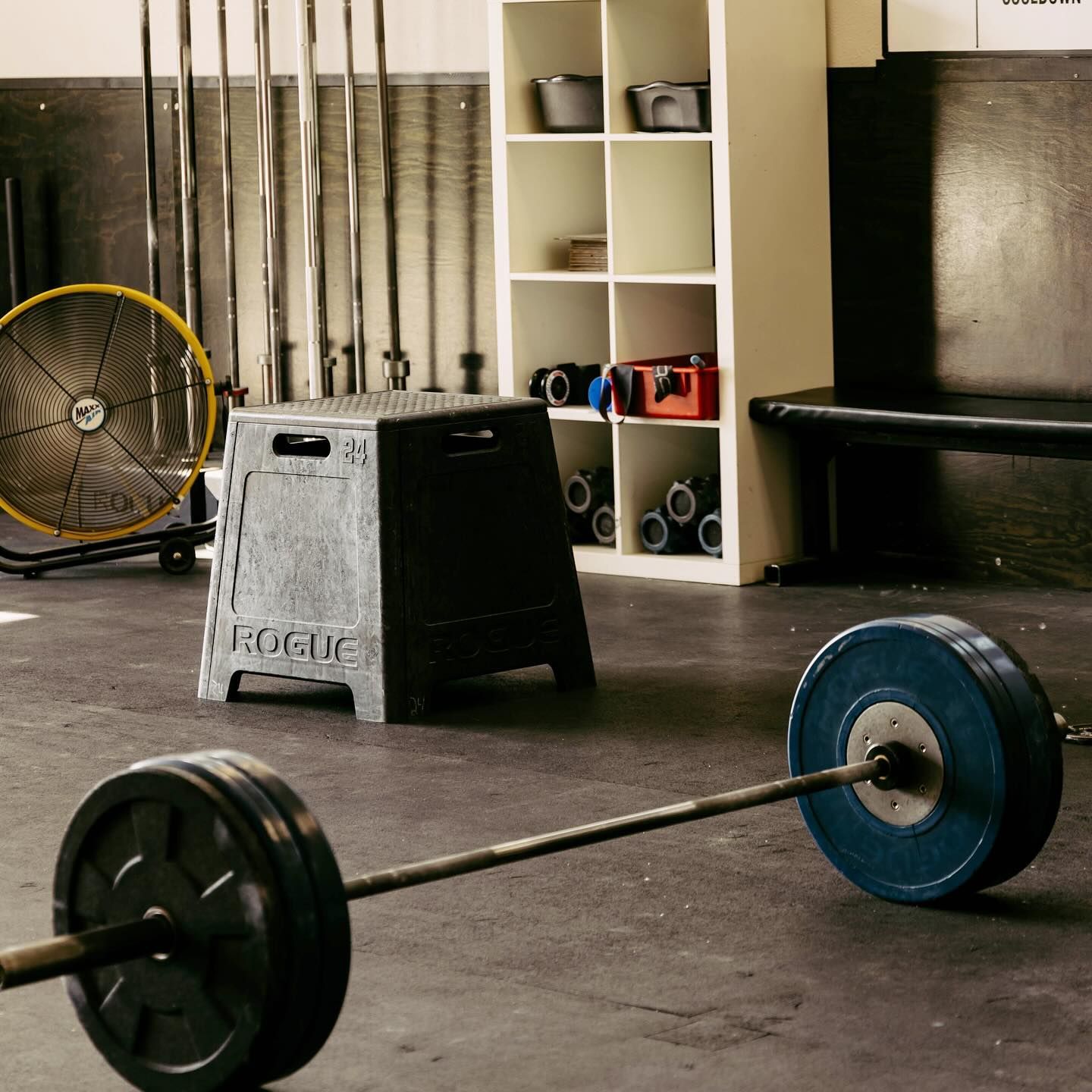 Barbell on floor in gym, Rogue step box, weights, fan, storage shelves, and weightlifting bars in the background.