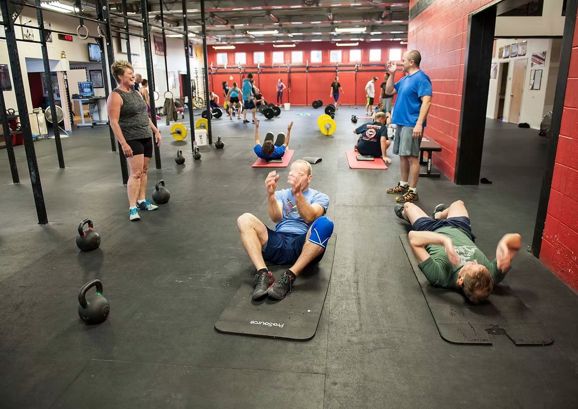 People exercising in a gym: floor exercises, kettlebells, red walls, and other gym-goers in the background.