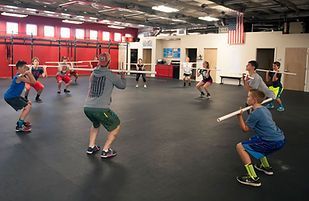 People in a gym squatting while holding sticks, led by a man. Red wall, U.S. flag visible.