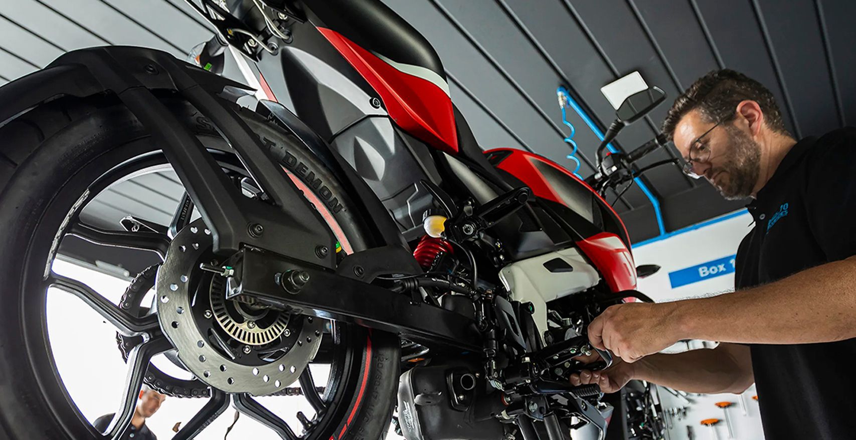 Mecânico trabalhando na parte inferior de uma motocicleta vermelha e preta em uma oficina.