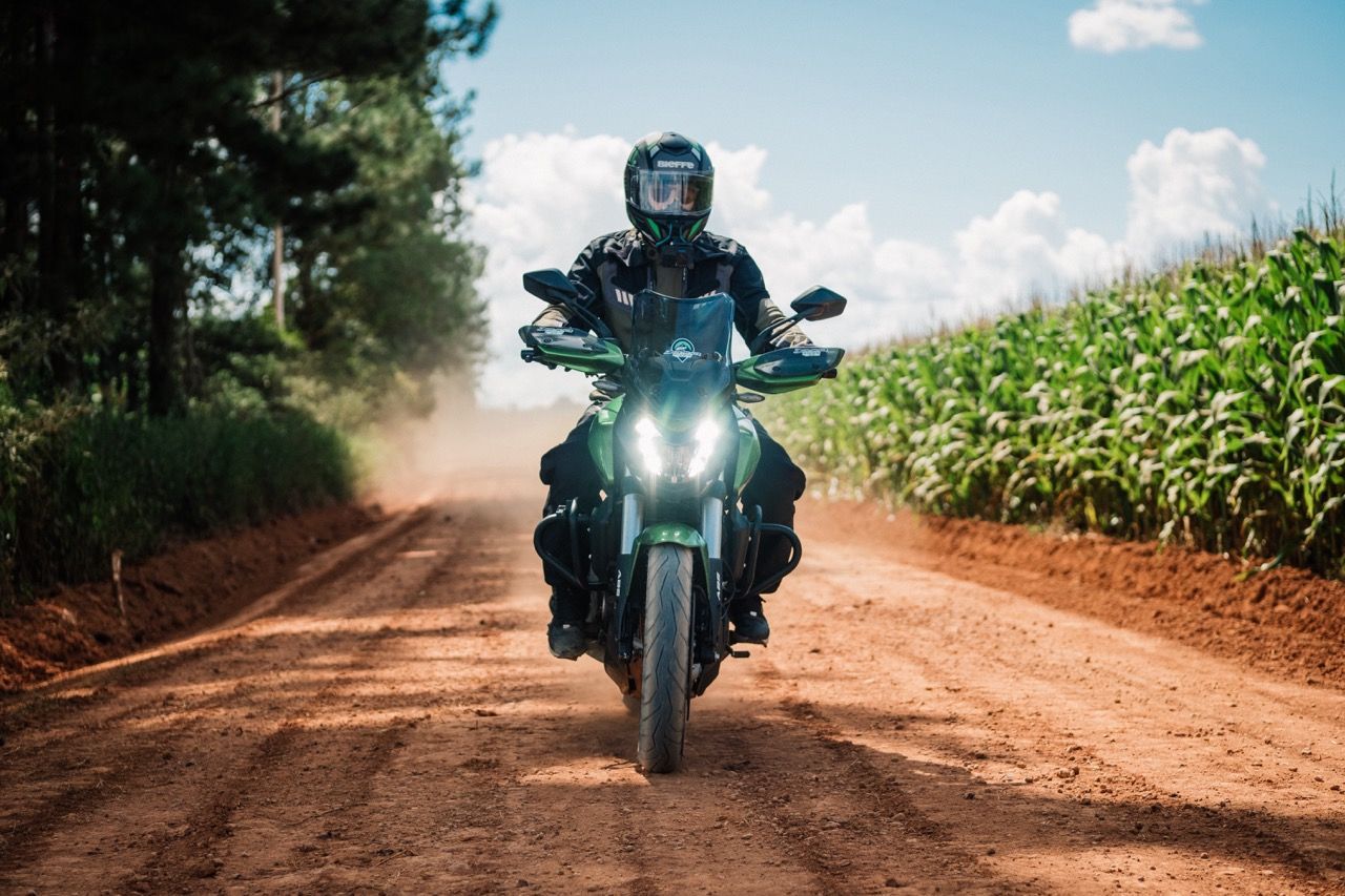 Motociclista andando em uma estrada de terra, com árvores e milharal ao fundo.