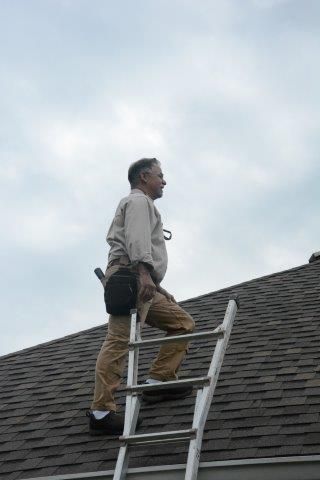 Man on a ladder inspecting a shingled roof, cloudy sky overhead.