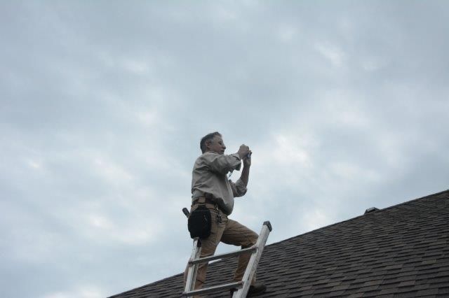 Man on a ladder taking a photo of the sky from a rooftop under a cloudy sky.