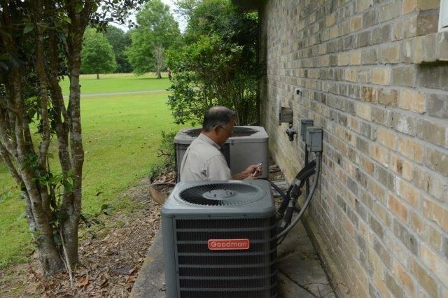 Man servicing an air conditioning unit next to a brick wall and a grassy yard.